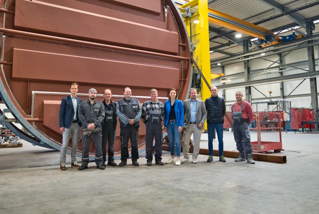 Gruppe von Menschen steht vor großer Industriemaschine in Fabrikhalle.