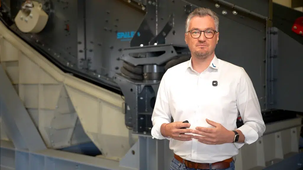 SPALECK screening machine expert in a white shirt standing in front of a dark-colored screening machine for bulky waste. The expert explains the advantages and machine settings of the screening machine, which is optimized for bulky waste.