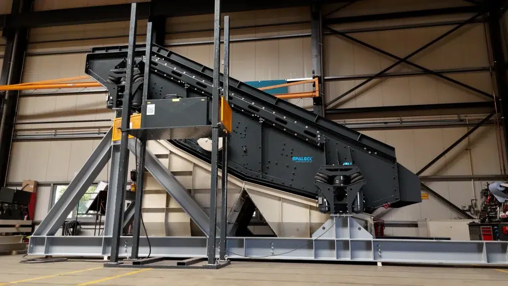 Dark-colored screening machine with the SPALECK logo and the SmartSUSPENSION air suspension system. The SPALECK recycling screen stands on a light gray substructure in the SPALECK screening machine production facility. Beneath the machine are two light beige fine-grain chutes, which will later, during ongoing operations at the recycling facility, discharge the screened material into two separate concrete bunkers. The screening machine is optimized for bulky waste.